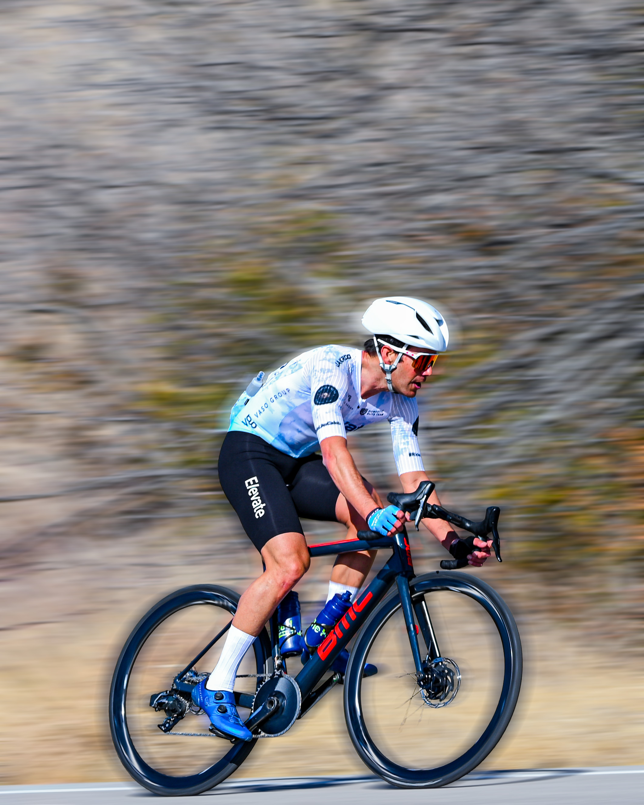 Cyclist wearing white helmet and blue cycling shoes rides a black and red BMC road bike on a paved road with blurred background.