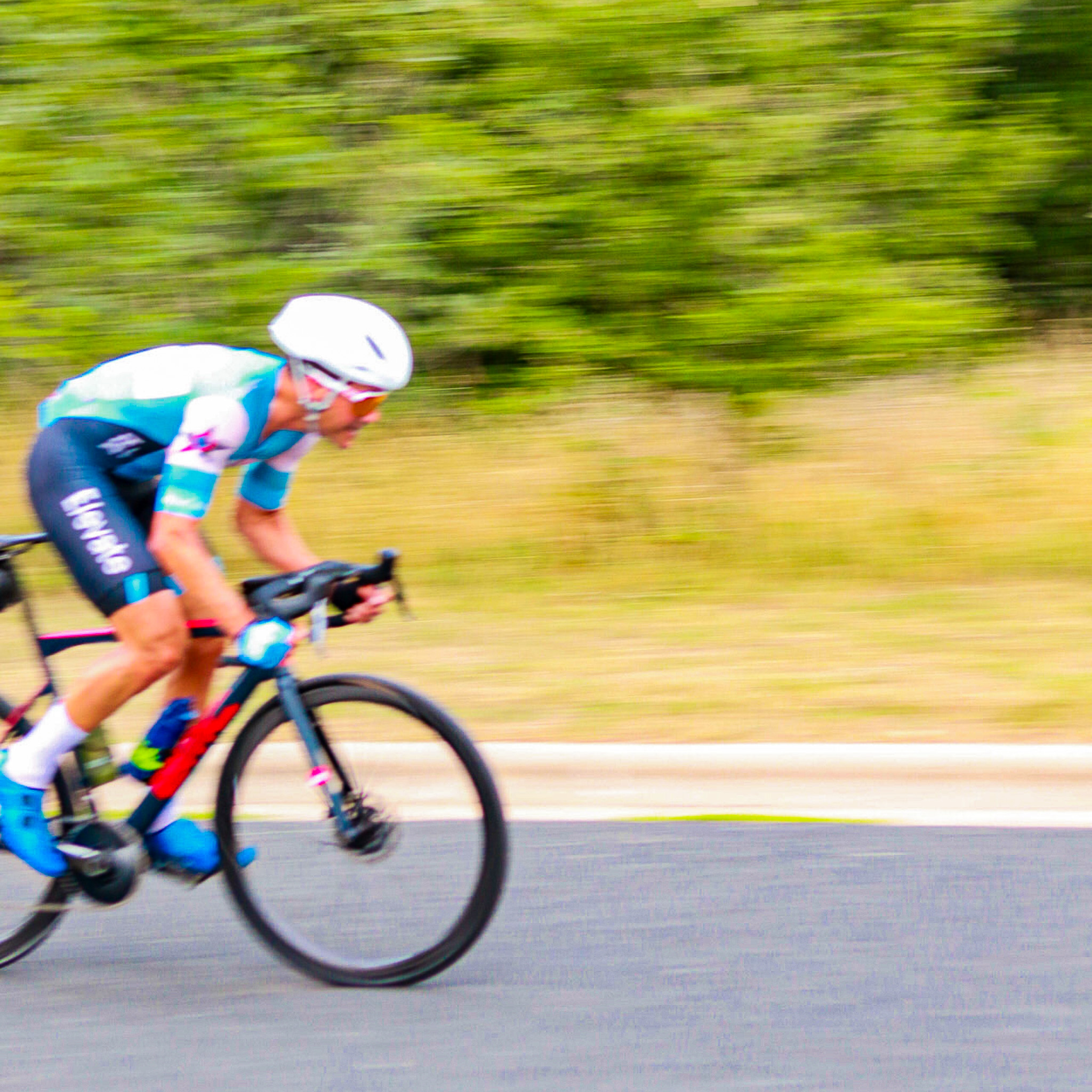 Cyclist in white helmet and blue cycling gear racing on a road with blurred background of greenery.