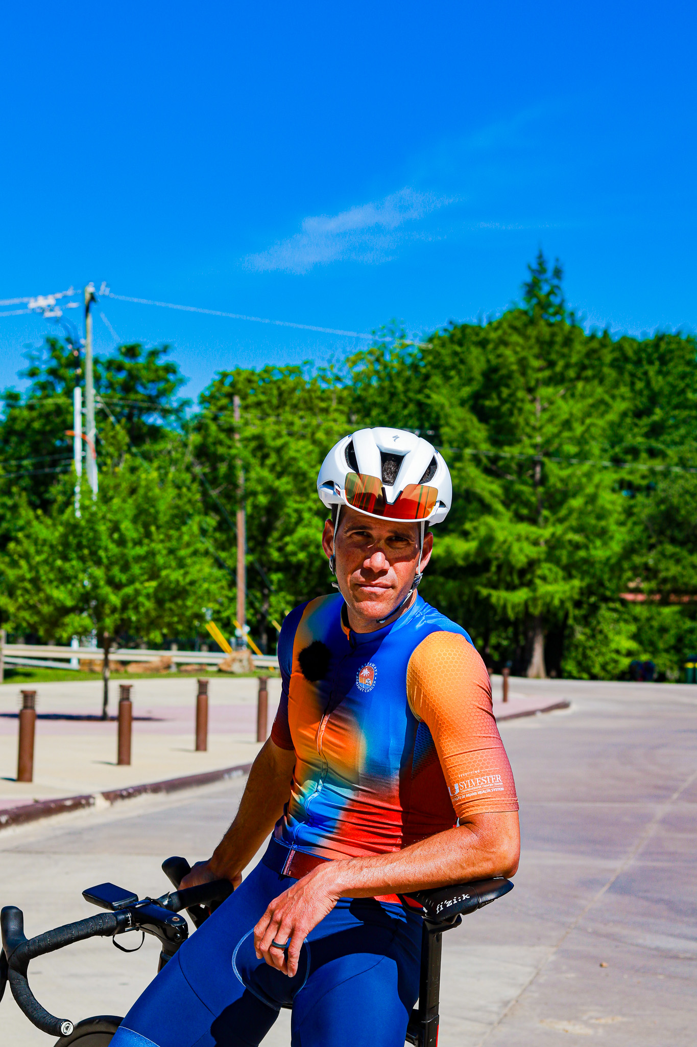 Male cyclist wearing a colorful blue and orange jersey and white helmet sitting on a bicycle outdoors on a sunny day.