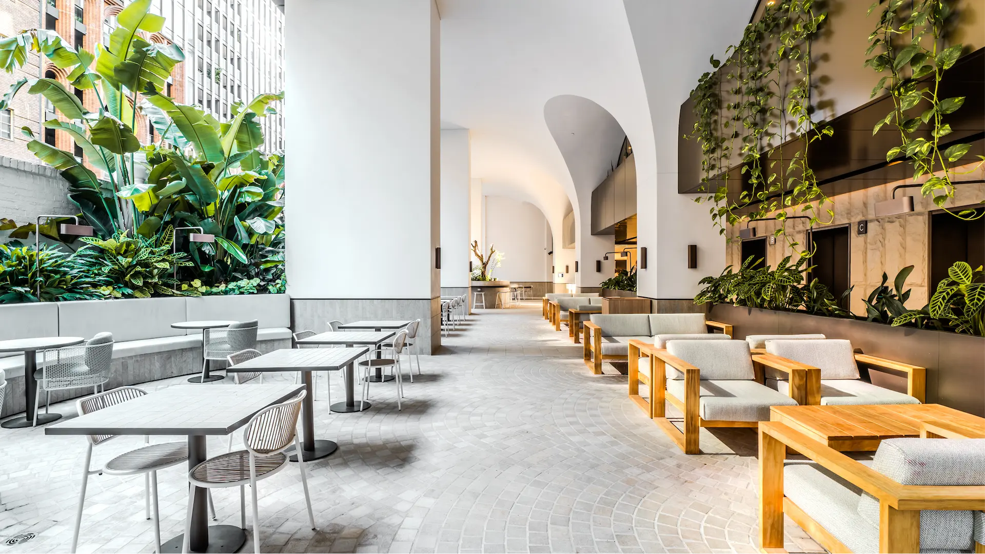 Bright modern indoor seating area with white tables and chairs on the left, and wooden framed sofas with cushions on the right, surrounded by lush green plants.