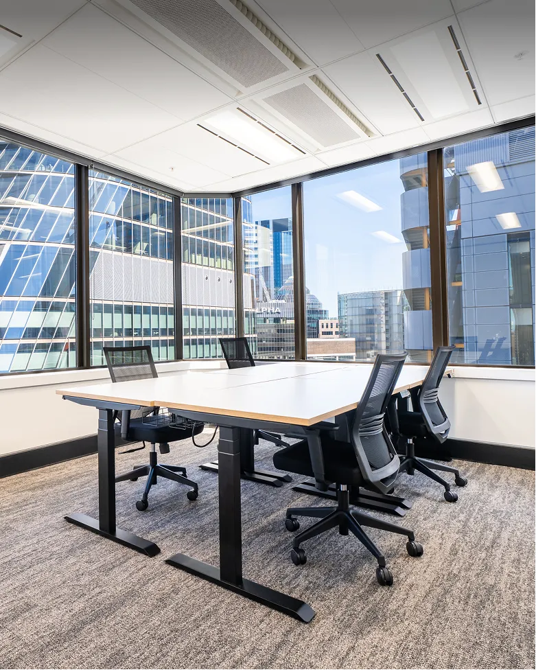 Modern office meeting room with four black mesh chairs around a rectangular table and large windows showing city buildings.
