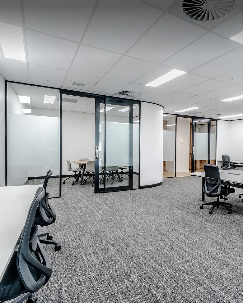 Modern office space with gray carpet, black office chairs, white desks, and glass-walled meeting rooms.