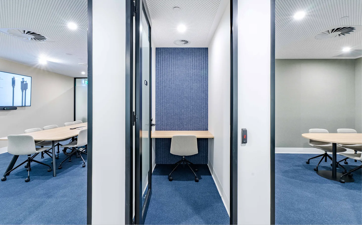 Modern office space with a small private cubicle containing a chair and desk, flanked by two glass-walled meeting rooms with tables and chairs.