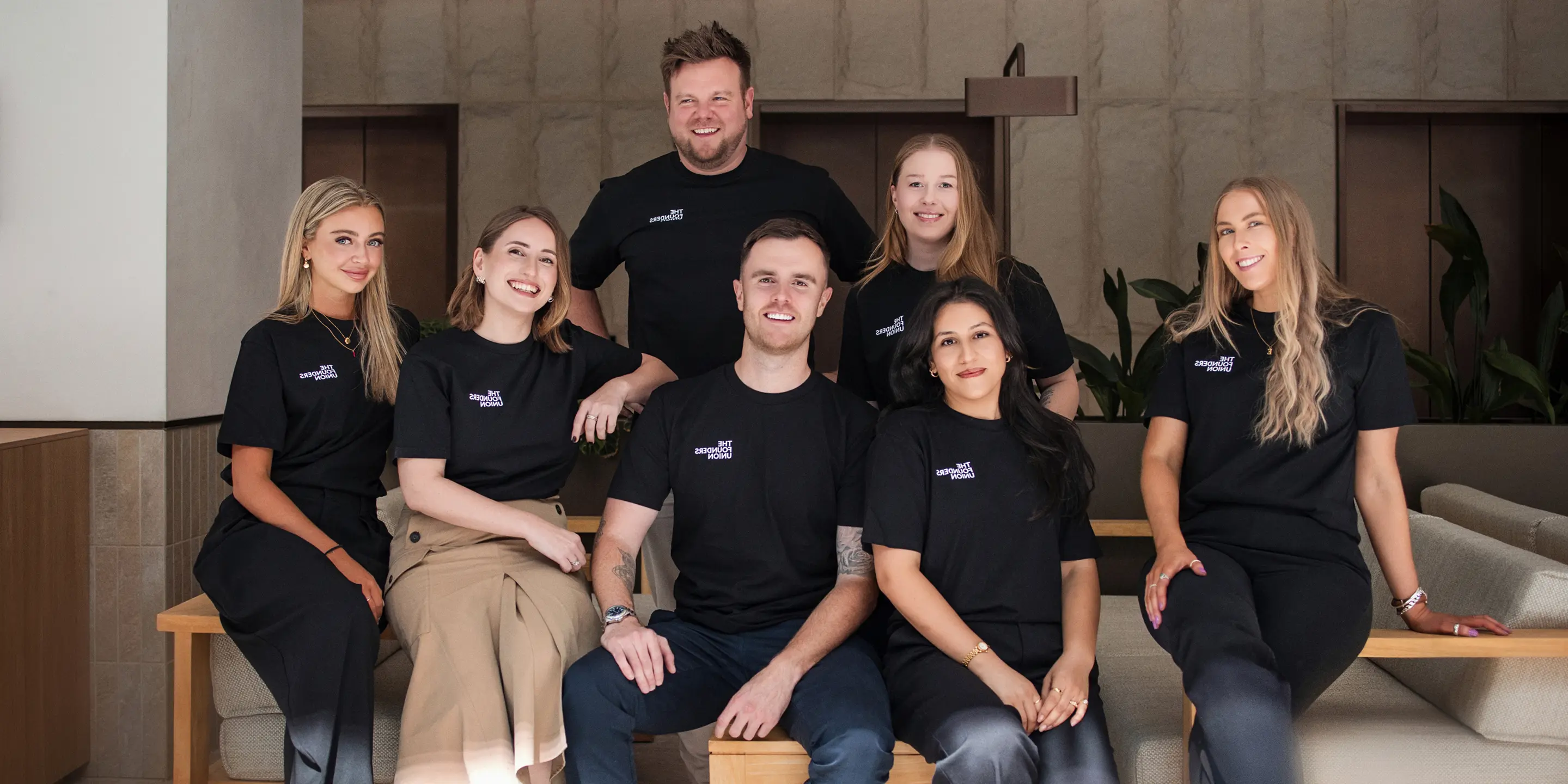 Group of seven young professionals wearing matching black t-shirts with white text, posing and smiling indoors on wooden benches with a modern background.