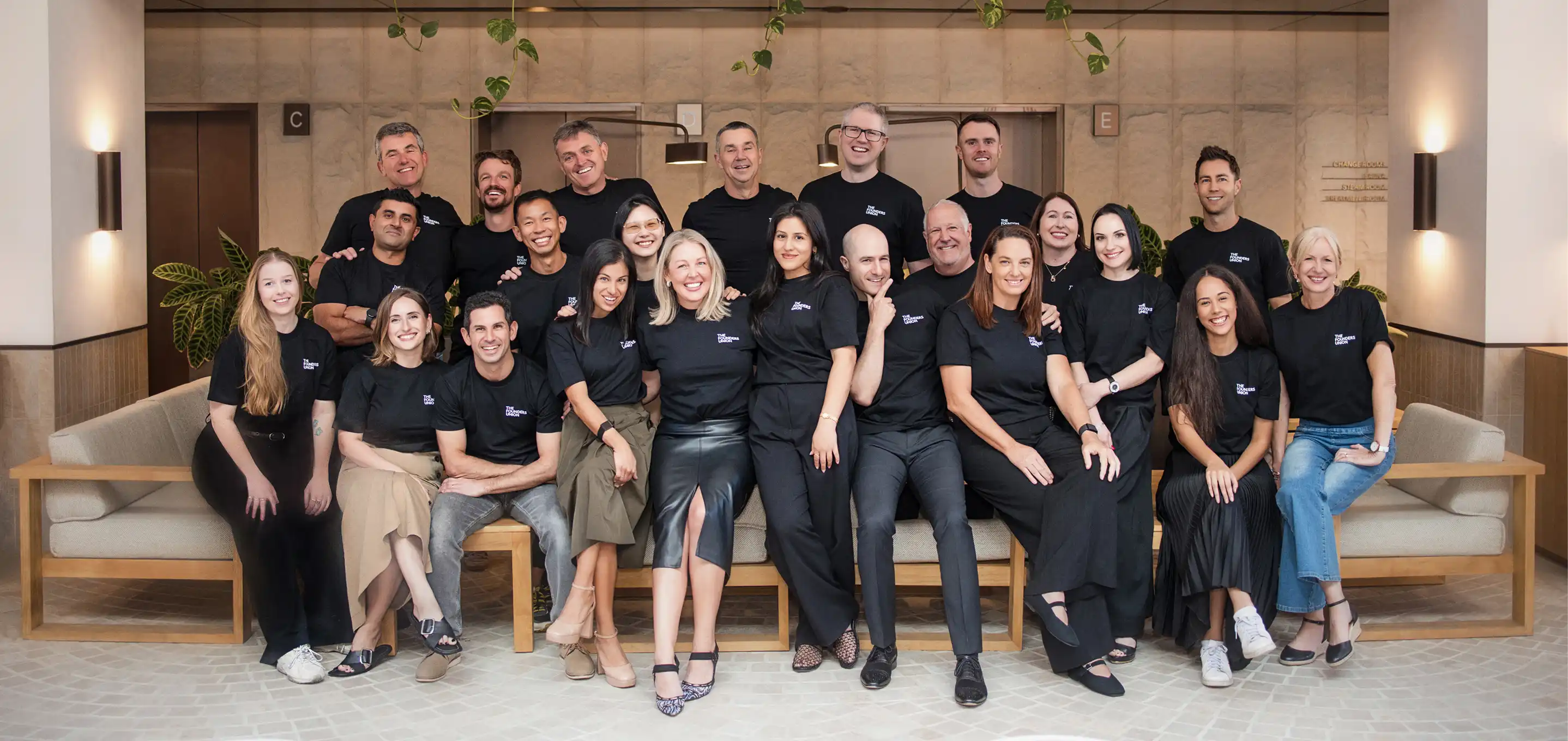 Group of twenty-two diverse men and women wearing matching black t-shirts with 'The Founders Nation' logo, posing and smiling in a modern indoor setting with couches and plants.