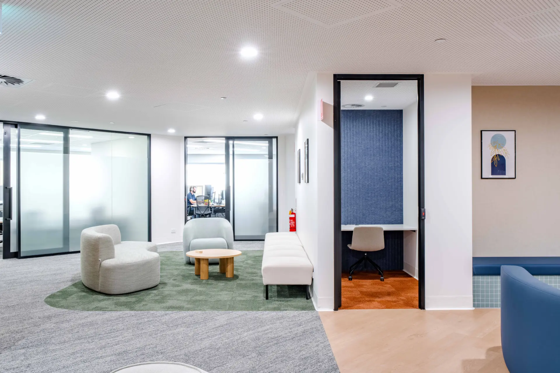 Modern office lounge area with gray chairs, a round wooden table on a green rug, a white bench, and a private workspace with a desk and chair.
