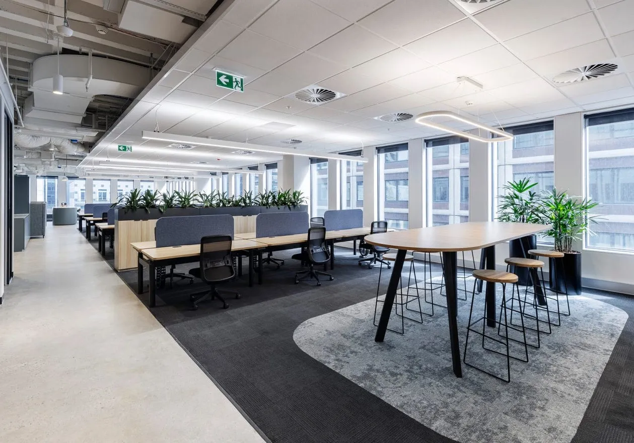 Modern open office space with rows of desks, chairs, and a large wooden table with stools near windows.