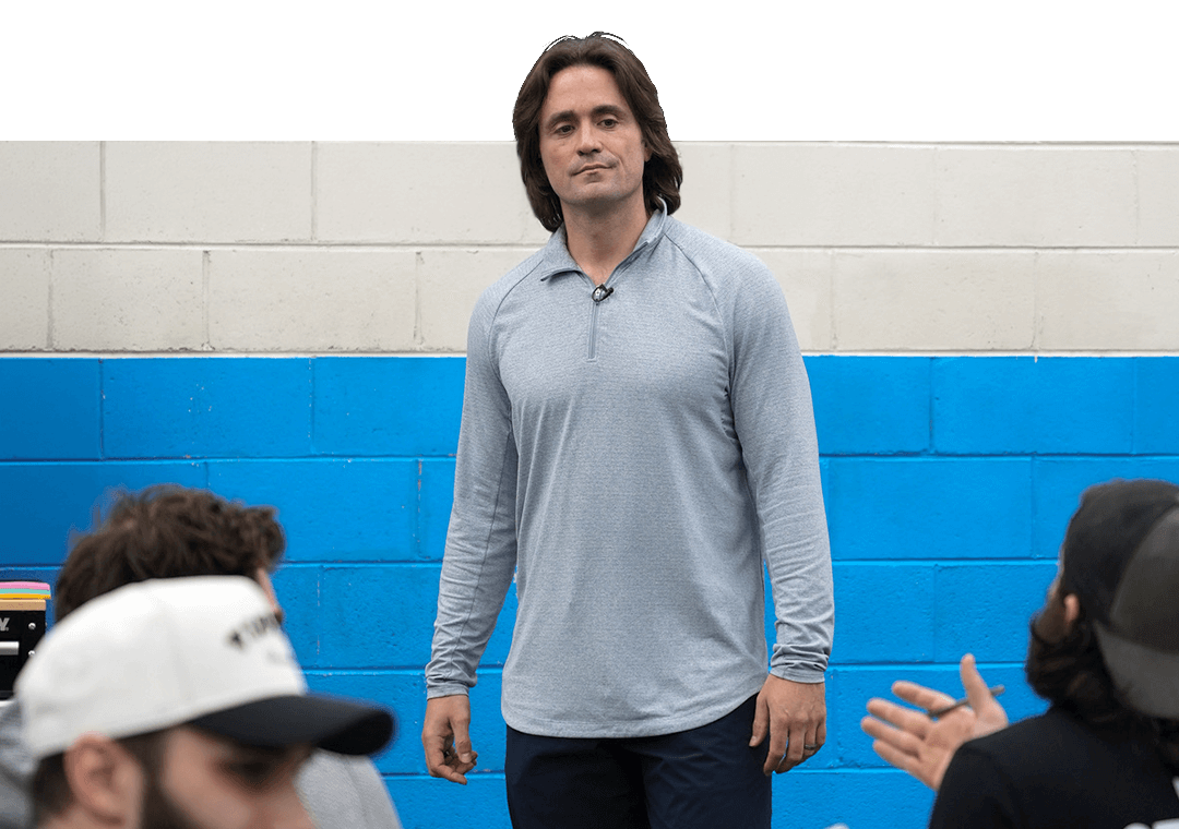Coach Vinny standing and speaking in front of a blue and white painted brick wall with people seated around him.