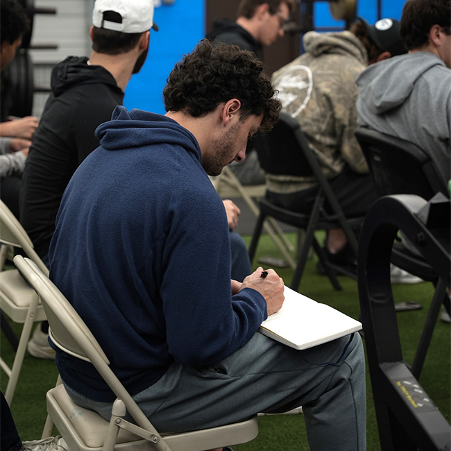Young man in a blue hoodie sitting on a folding chair, writing in a notebook during a group class or workshop.