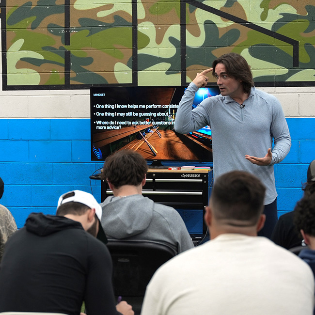 Coach Vinny giving a presentation to a seated audience with a TV screen showing bullet points about mindset.