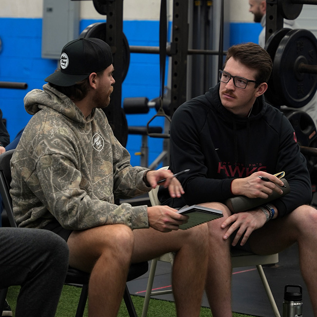 Two young men sitting and conversing in a gym with weightlifting equipment in the background, one holding a notebook and pen.