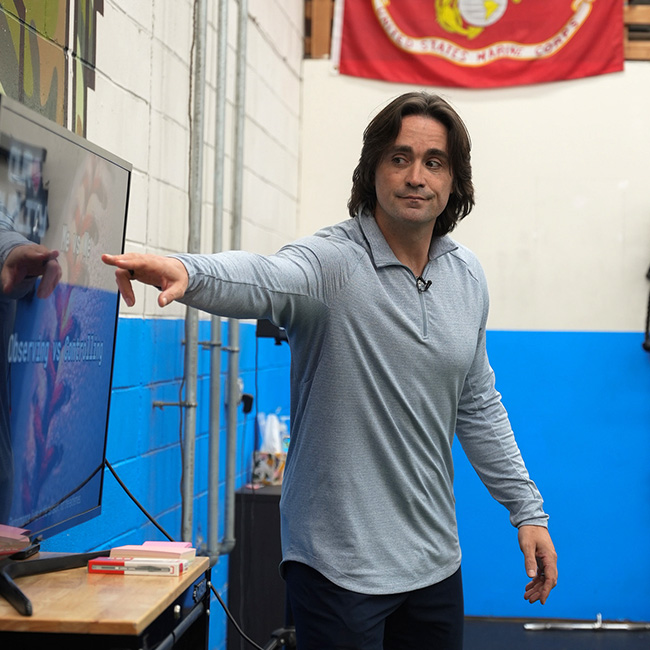 Coach Vinny pointing at a screen in a gym with blue and white walls and a Marine Corps flag in the background.
