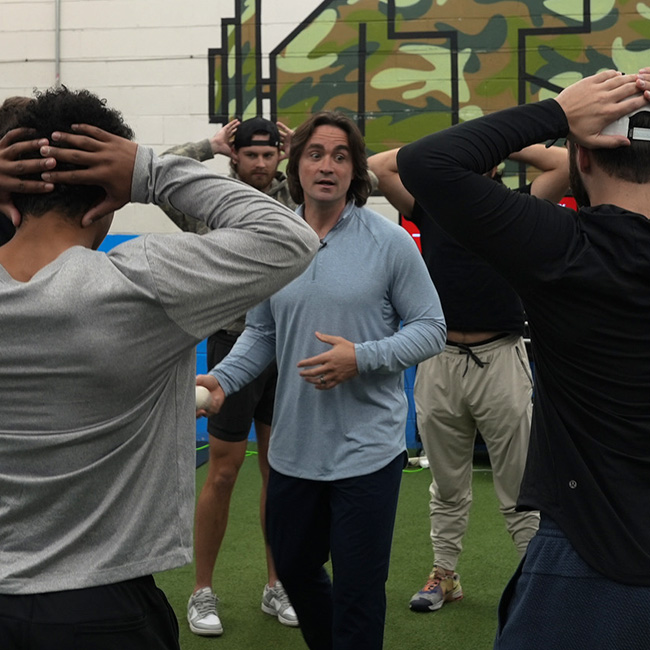 Coach Vinny in blue long-sleeve shirt demonstrating an exercise to a group of men with hands on their heads in an indoor gym.