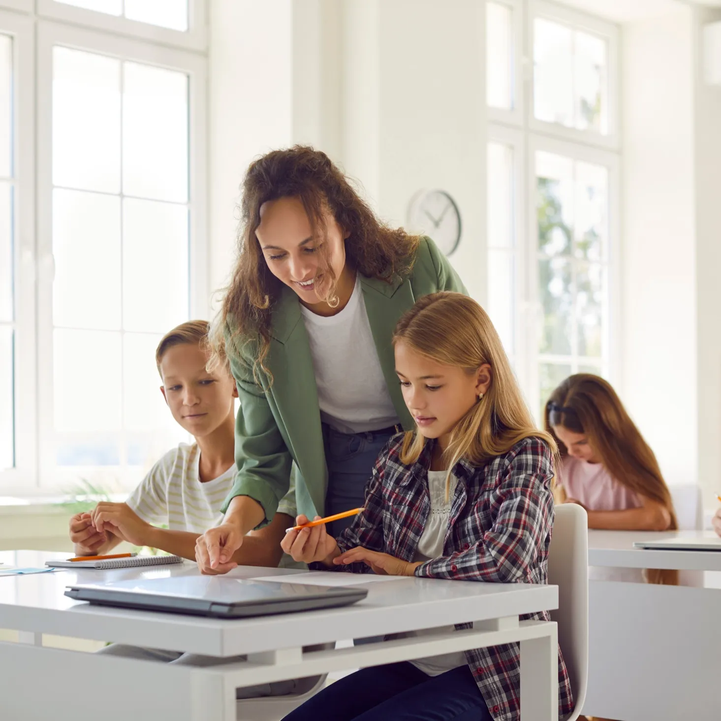 Teacher assisting two students with schoolwork at a bright classroom desk with a laptop.