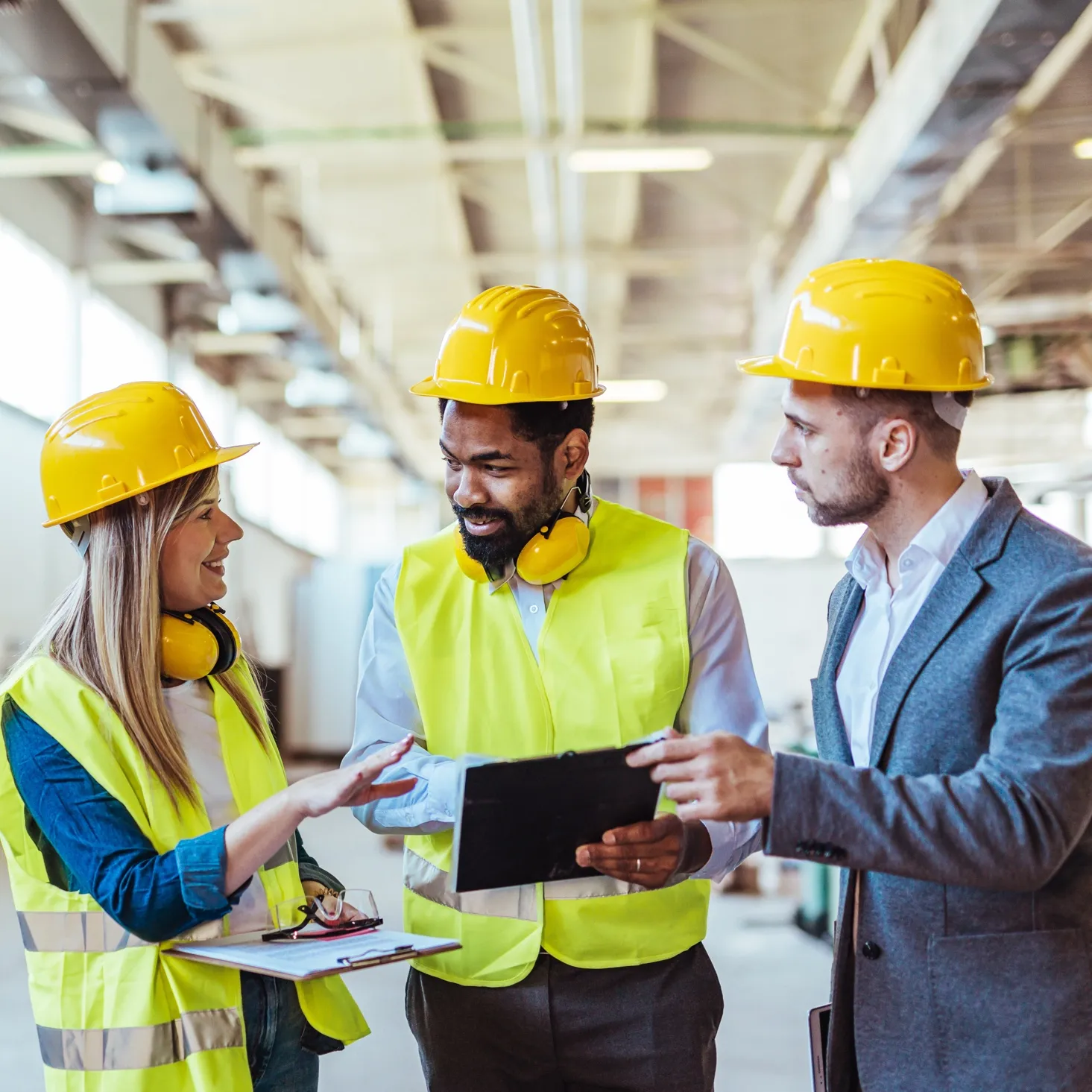 Three construction professionals wearing yellow hard hats and safety vests discussing documents inside a building under construction.