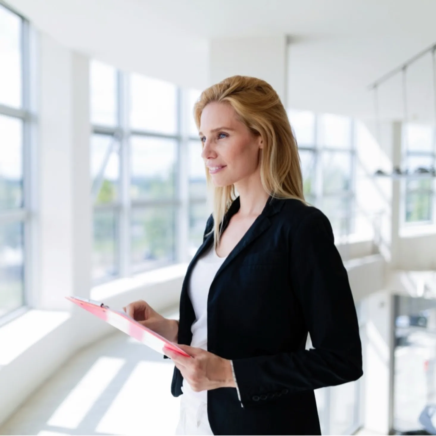 Smiling blonde woman in black blazer holding a pink clipboard, standing in a bright office space with large windows.