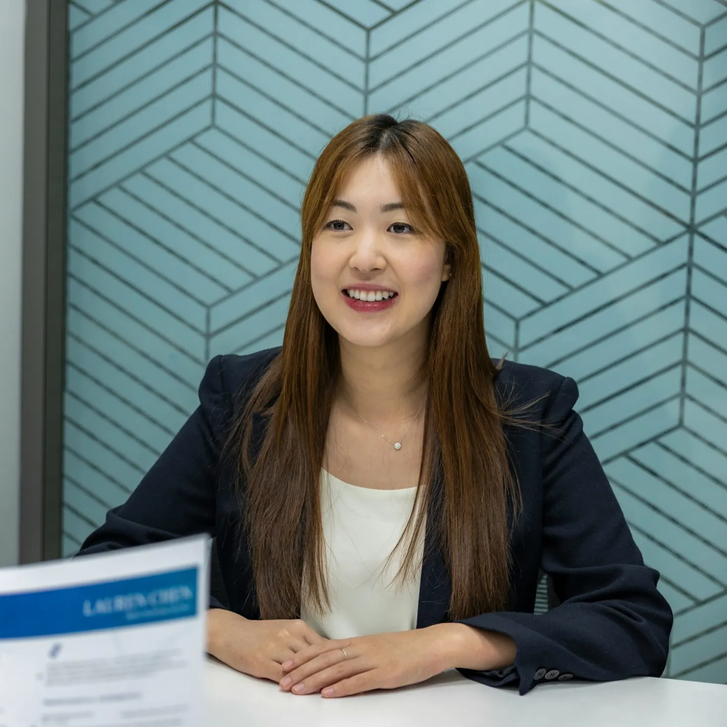 Smiling young woman in business attire sitting at a table with a document in front of her.