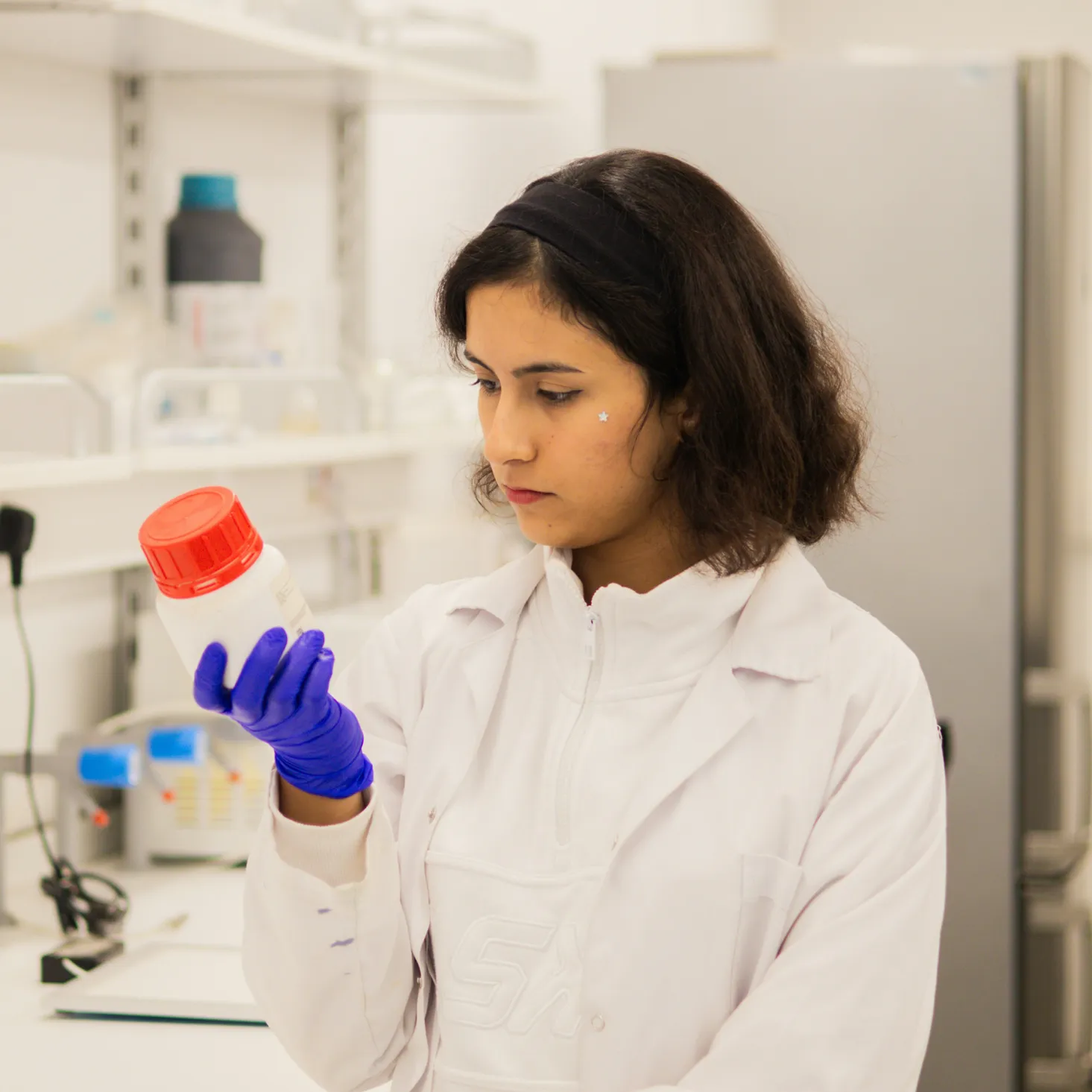 Scientist with dark hair and blue gloves examining a white laboratory bottle with a red cap in a lab setting.
