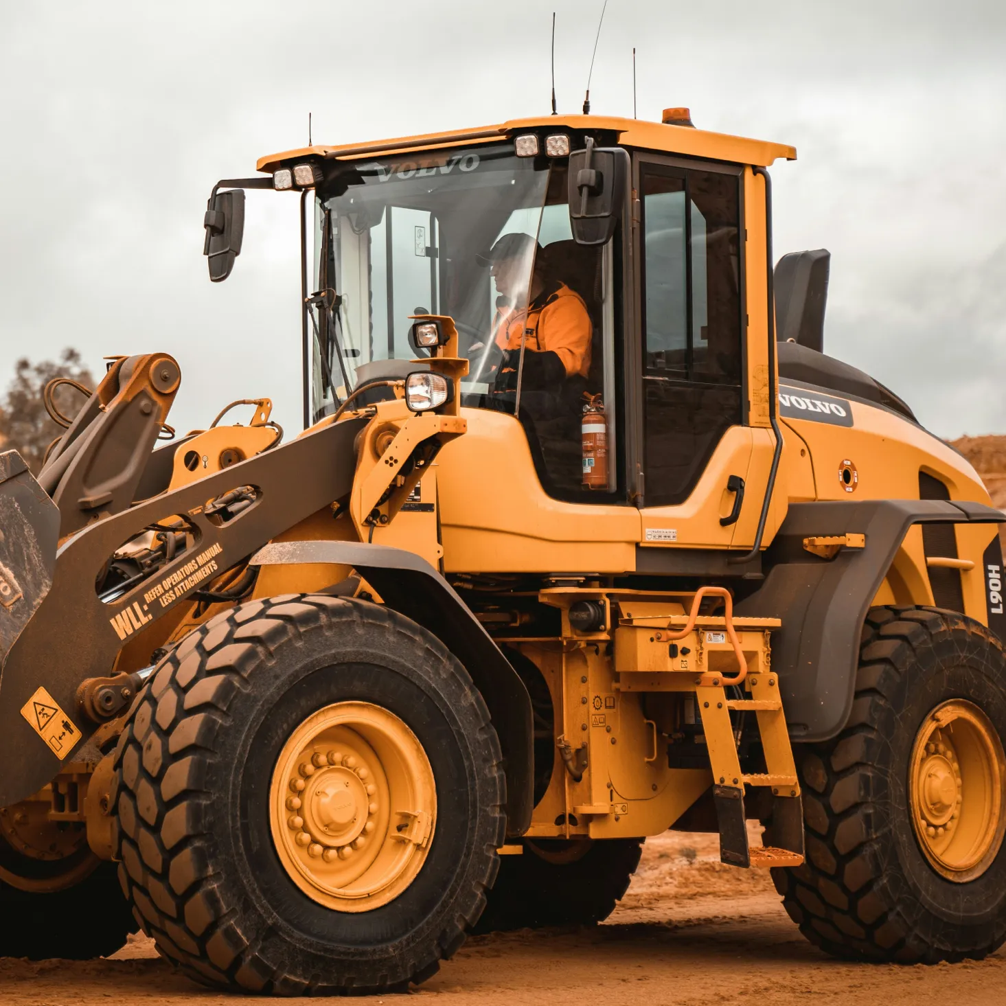 Operator in orange jacket driving a yellow Volvo front loader on a dirt site.