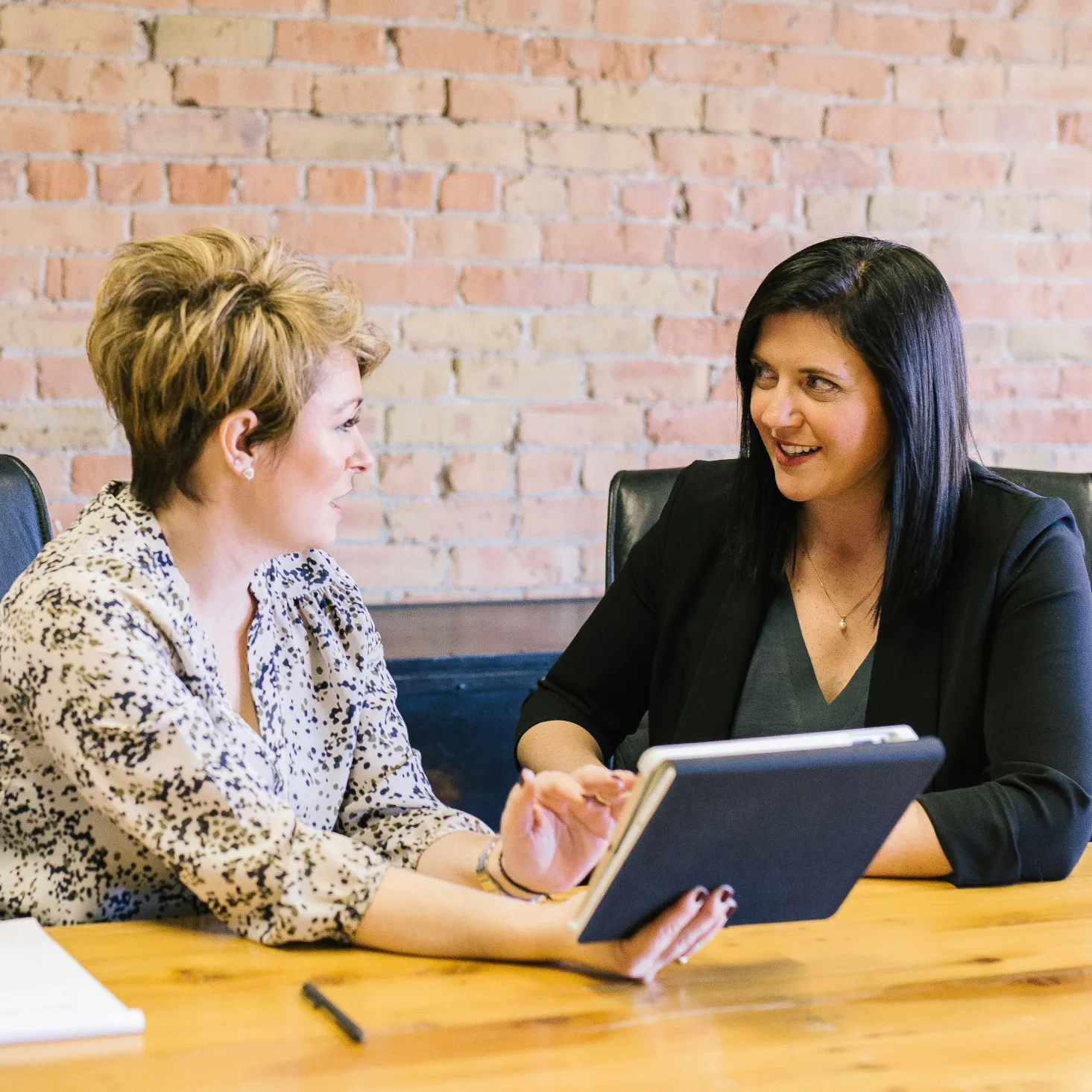 Two women in a meeting room discussing while one holds a tablet in front of the other.