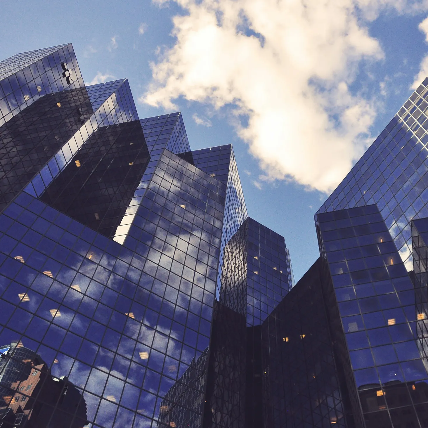 Modern glass skyscrapers reflecting clouds and blue sky from a low perspective.