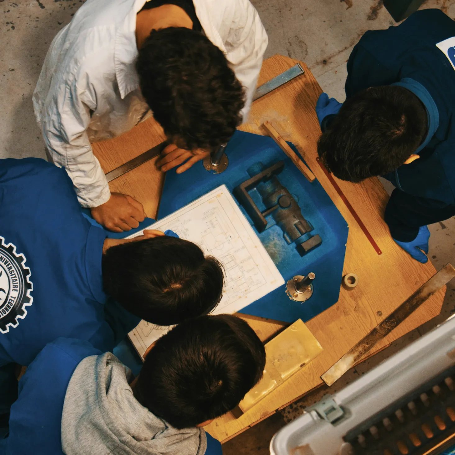 Top view of four people examining a technical drawing and a mechanical part on a wooden table.