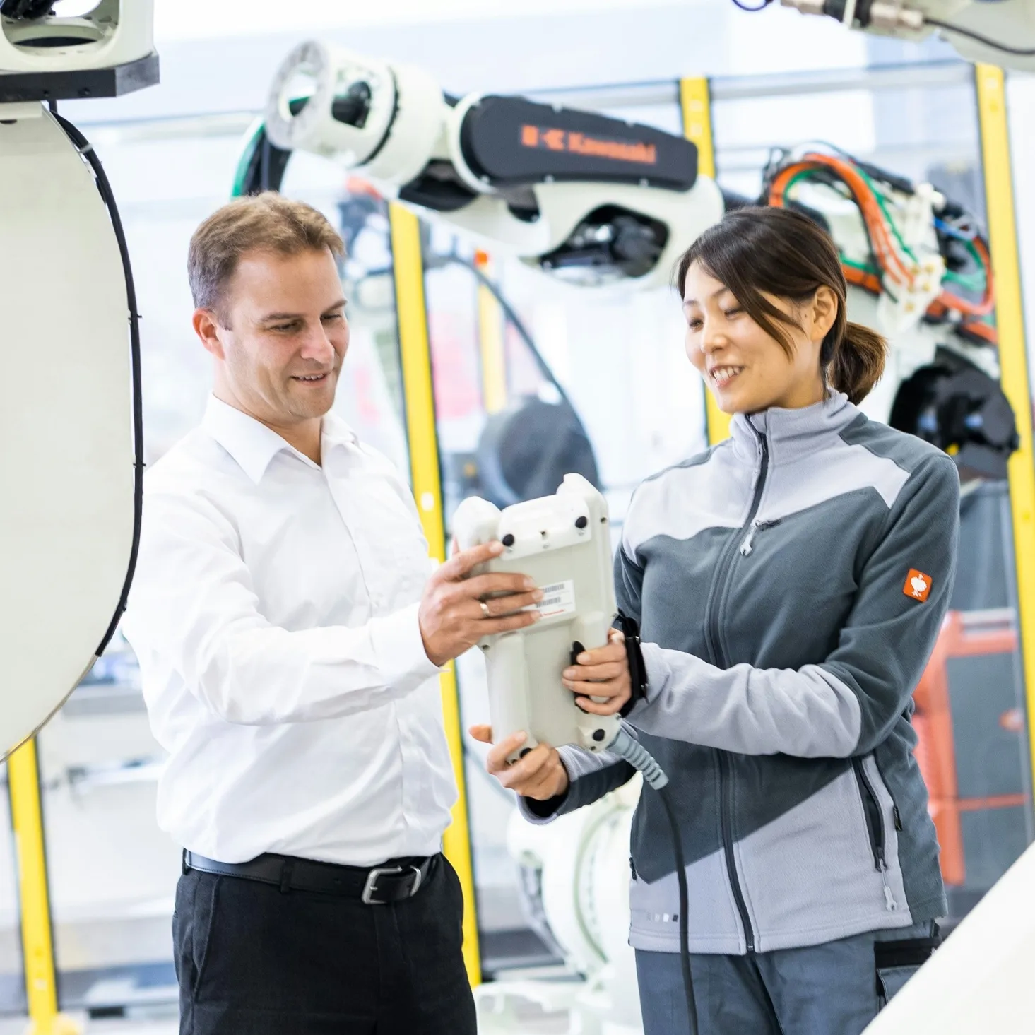 Two factory workers examining a robotic device in an industrial setting with robotic arms in the background.