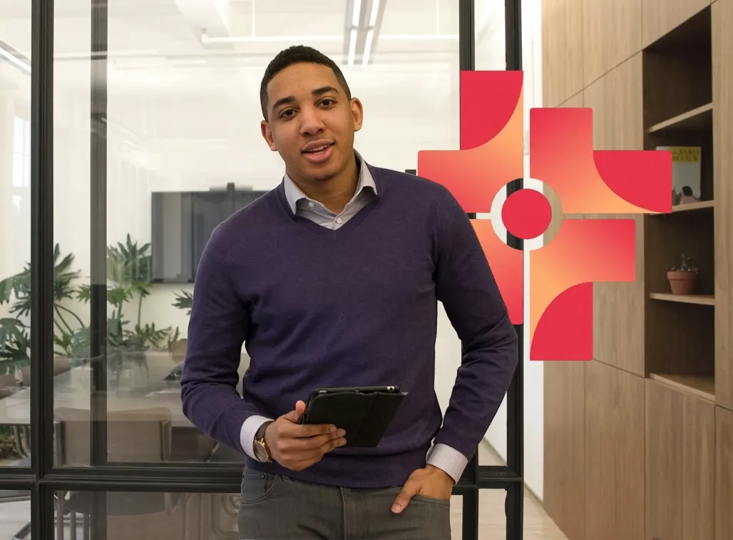 Young man in a purple sweater standing in an office holding a tablet with one hand in his pocket.