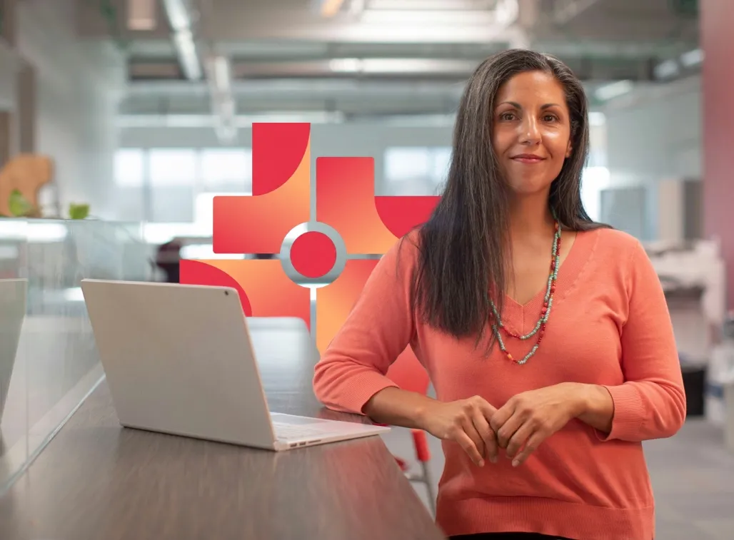 Smiling woman with long dark hair wearing an orange sweater and beaded necklace standing by a desk with a laptop in a modern office.