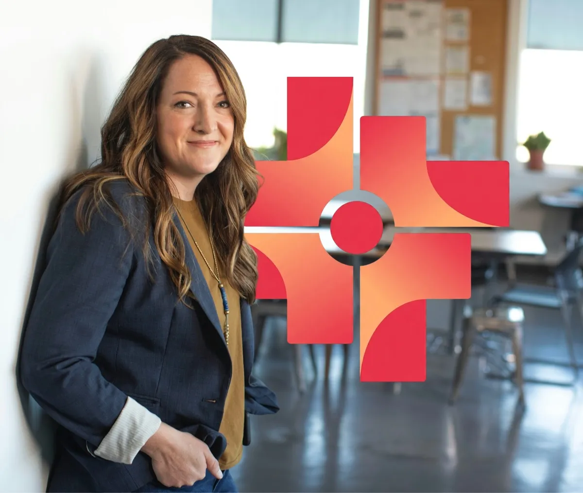 Smiling woman with long wavy hair wearing a blue blazer and mustard shirt leaning against a wall in a classroom setting with a red and orange abstract cross symbol overlaid.