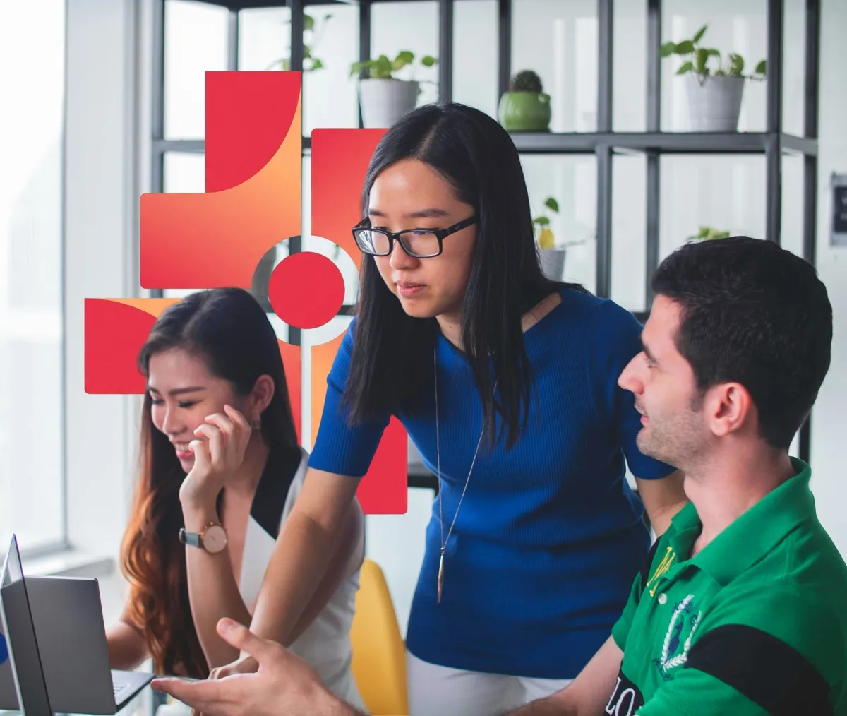 Three colleagues collaborating at a desk with a laptop, surrounded by potted plants on shelves.