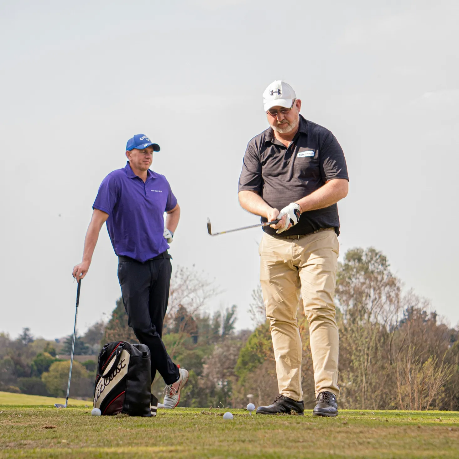 Two men on a golf course, one preparing to swing a golf club while the other leans on a golf club watching.