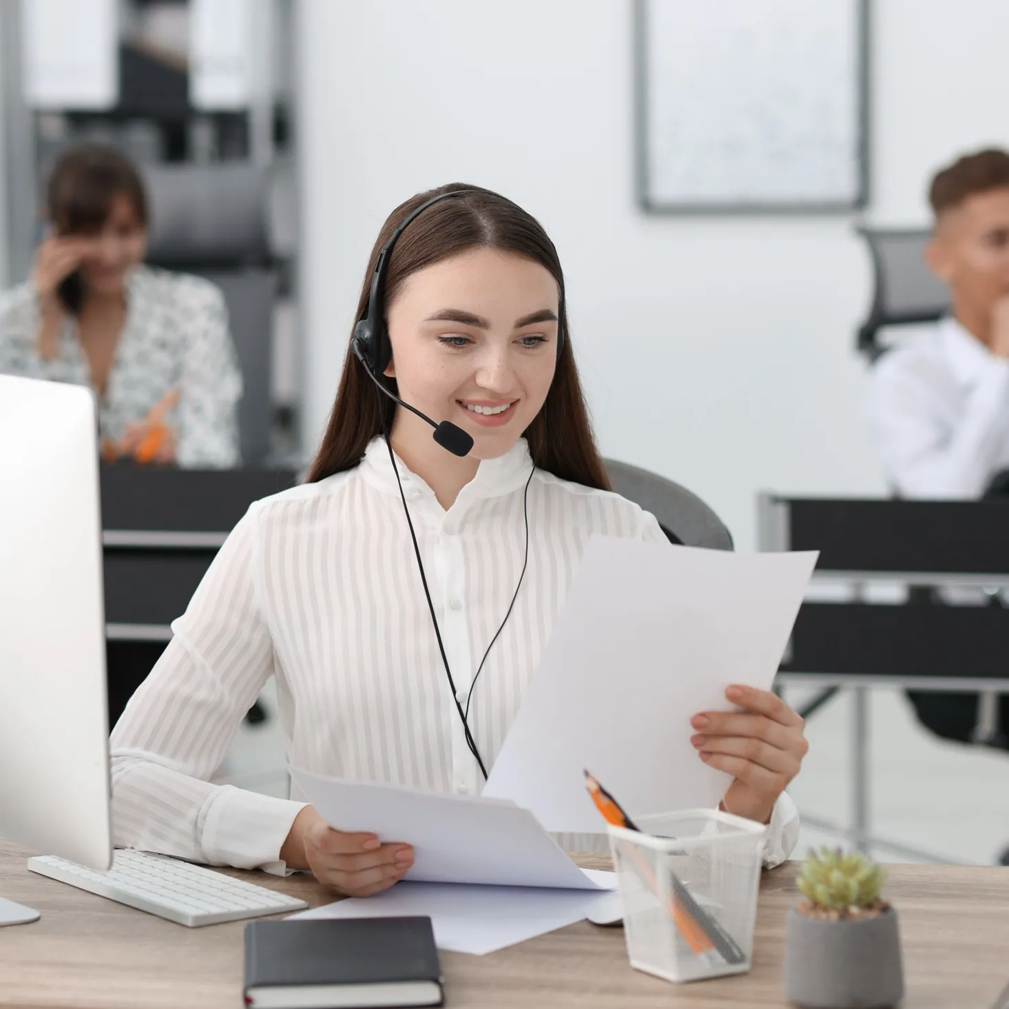 Smiling woman wearing a headset reviewing papers at her desk in a modern office.