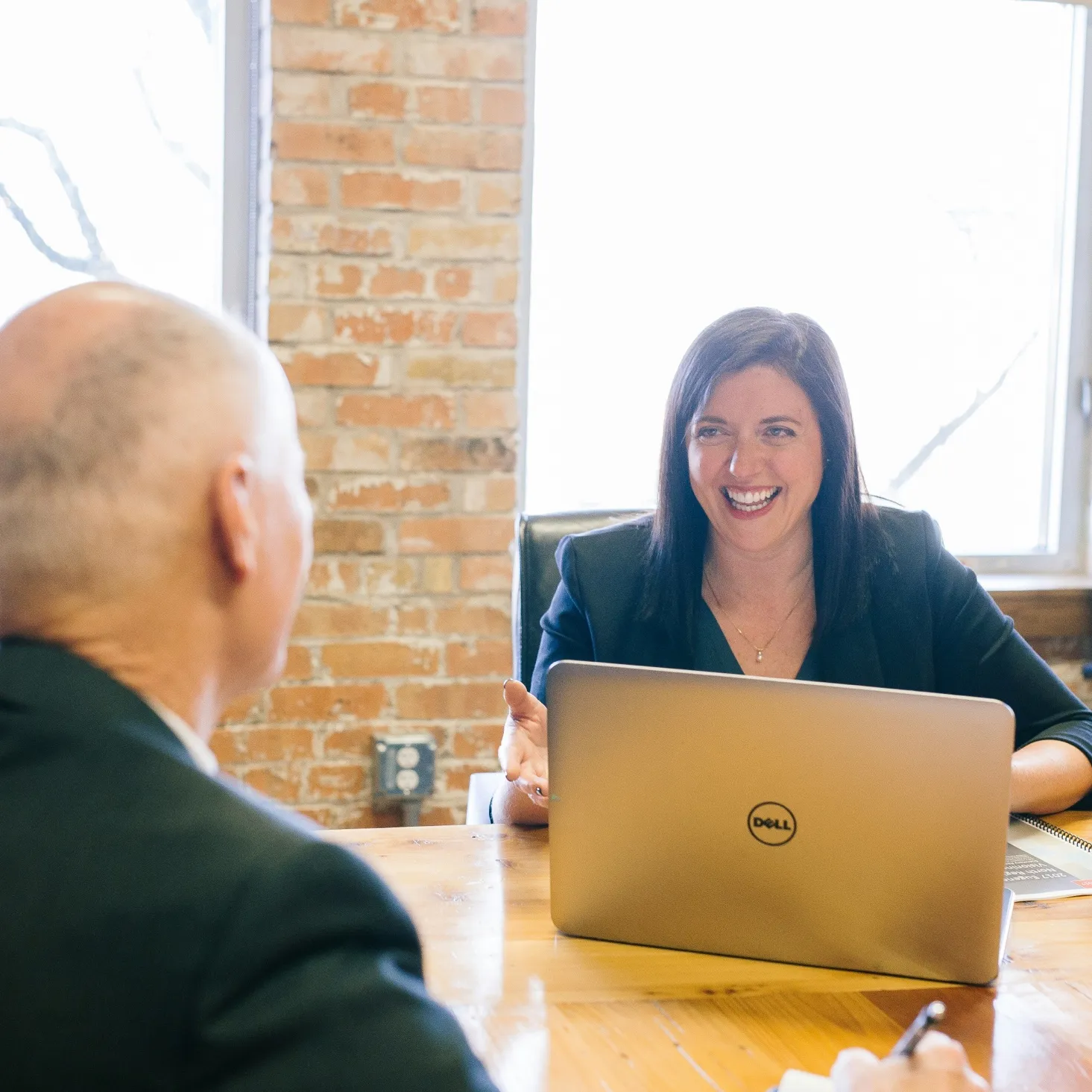 Smiling woman sitting at a wooden table with a laptop, talking to a man in a suit, in a room with a brick wall and large window.