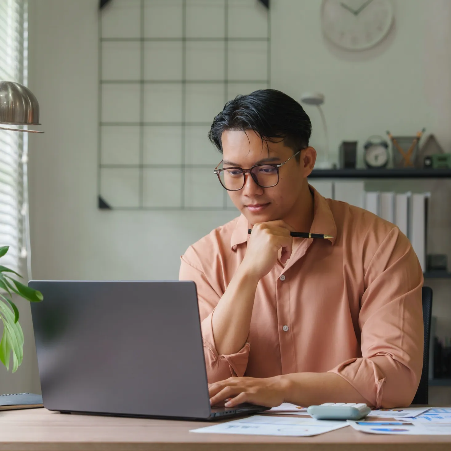 Man wearing glasses and a salmon shirt working on a laptop at a desk with papers and a calculator.