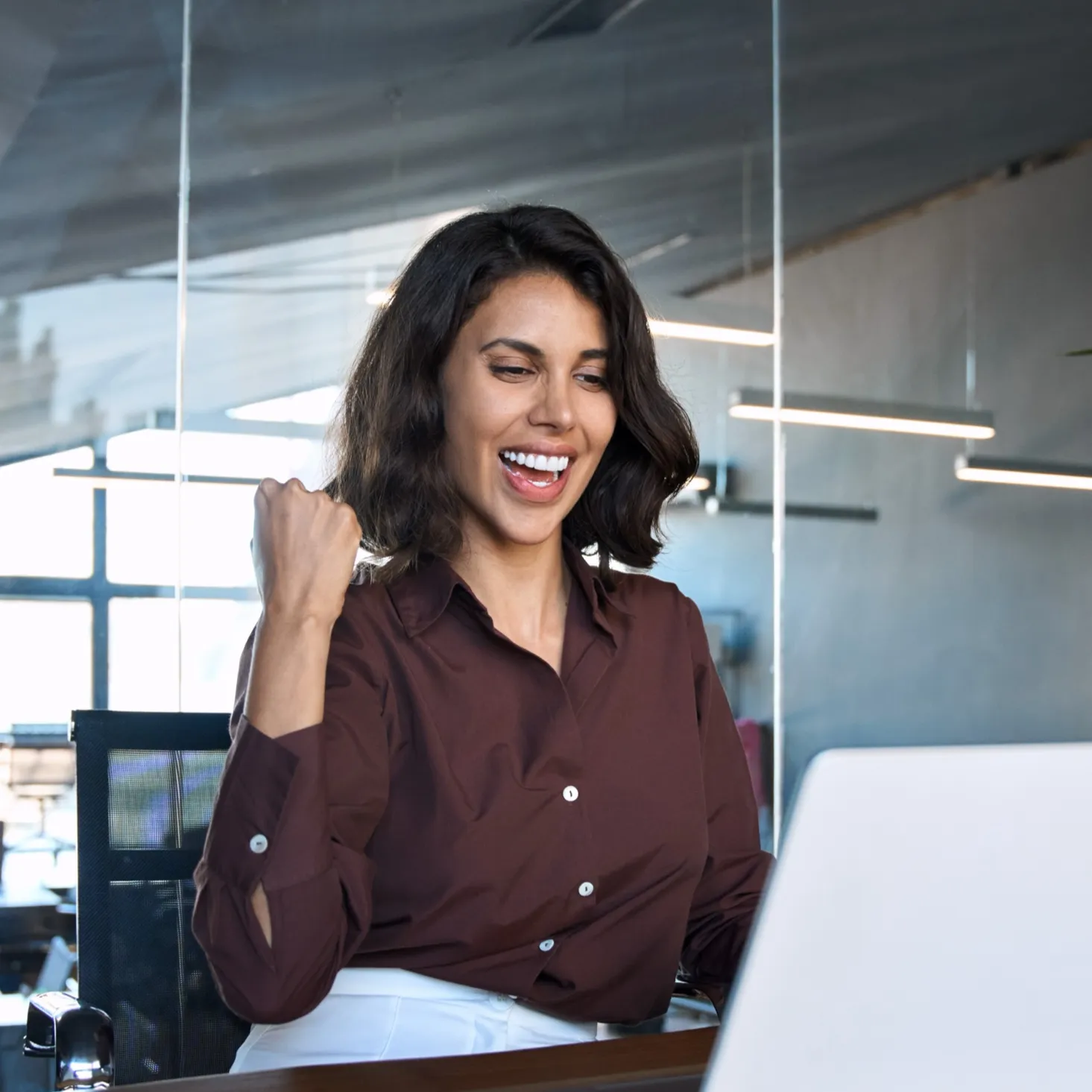 Smiling woman in a brown shirt sitting at a desk, looking at a laptop and raising her fist in a gesture of success.