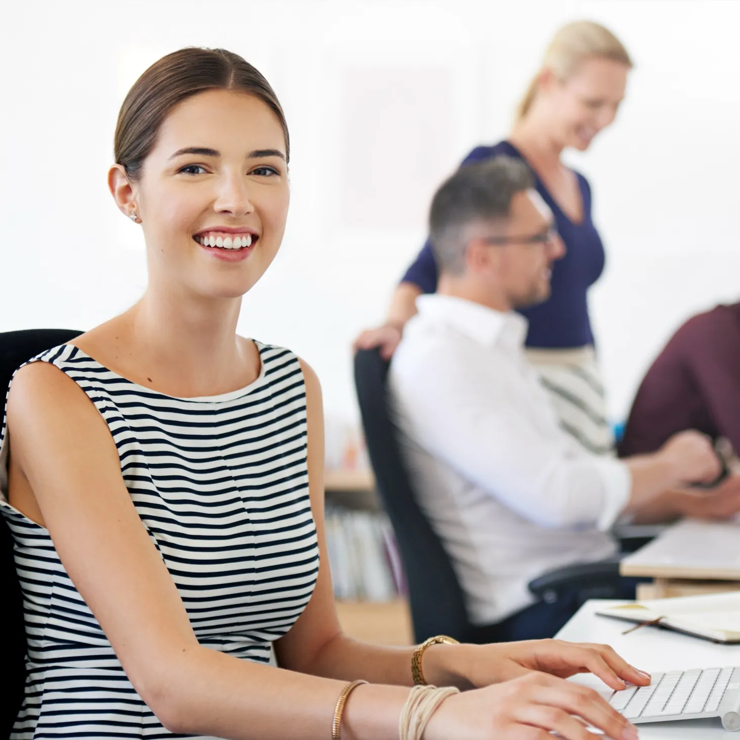 Smiling woman in a striped sleeveless top typing on a keyboard in an office with two blurred colleagues in the background.