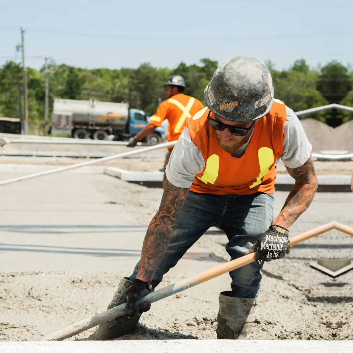Construction worker wearing an orange safety vest and helmet smoothing wet concrete with a tool on a sunny day.