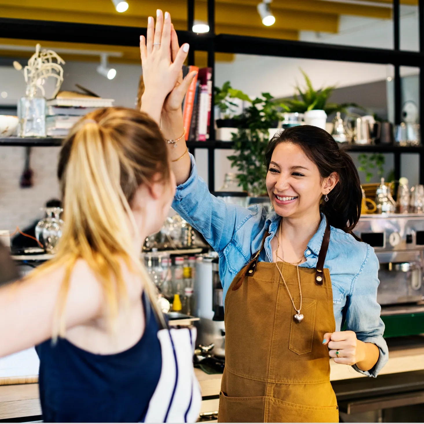 Two women in aprons giving a high five in a coffee shop kitchen.