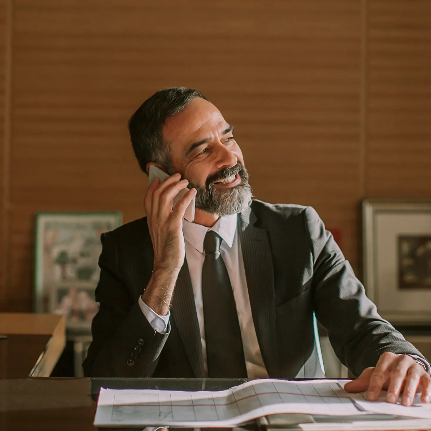 Smiling man in suit talking on phone while sitting at desk with open documents.