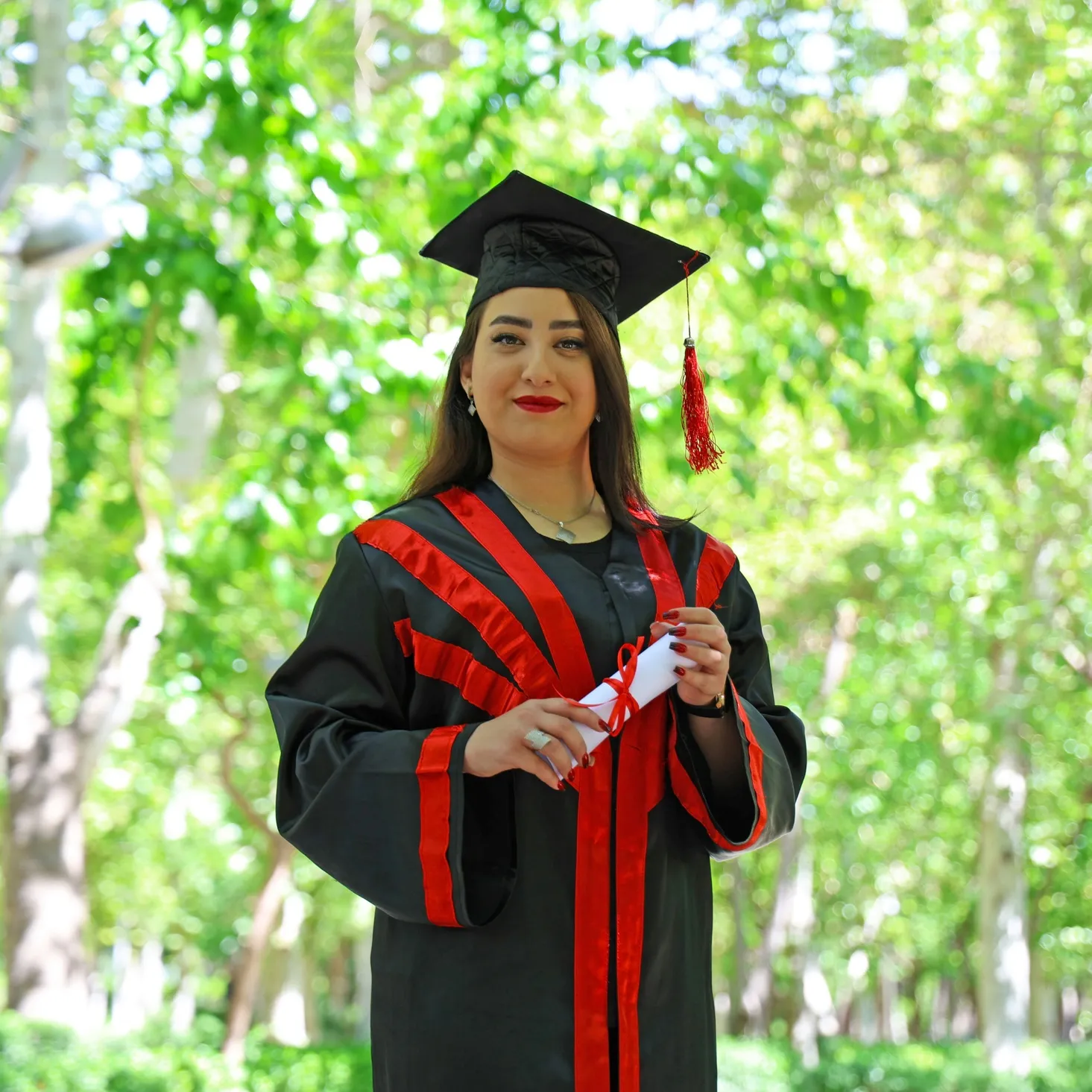 Smiling graduate woman in black and red academic gown and cap holding a diploma outdoors with green trees in the background.