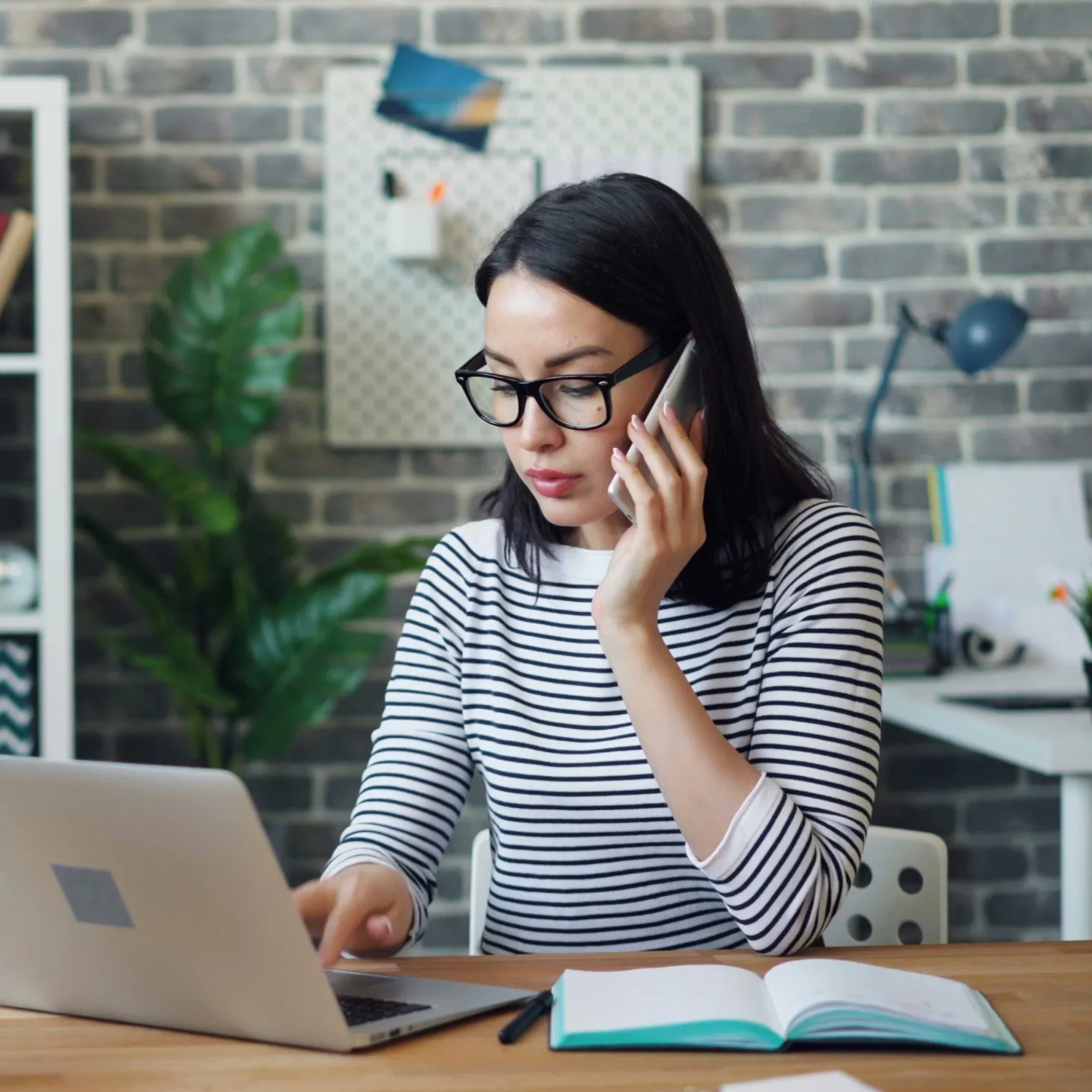 Young woman wearing glasses talking on a mobile phone while using a laptop at a wooden desk with an open notebook.