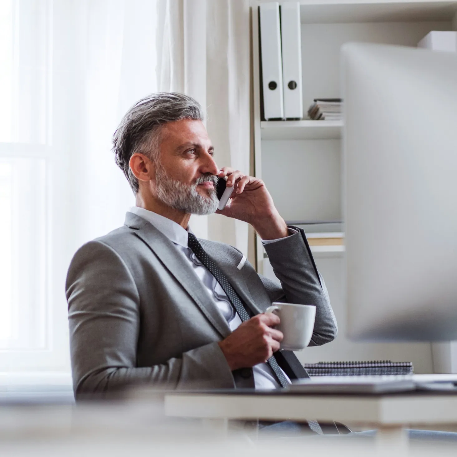 Middle-aged man with gray hair and beard in a gray suit talking on a phone while holding a white coffee cup at his desk.