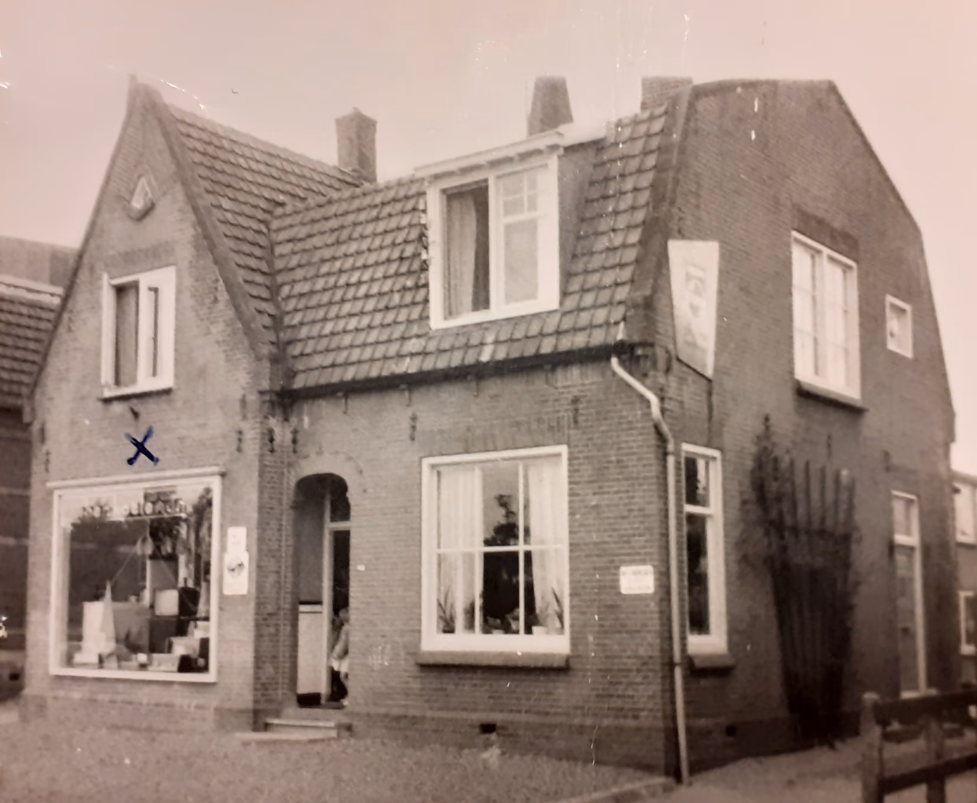 Oud zwart-witfoto van een stenen huis met pannendak en een winkel aan de voorkant.