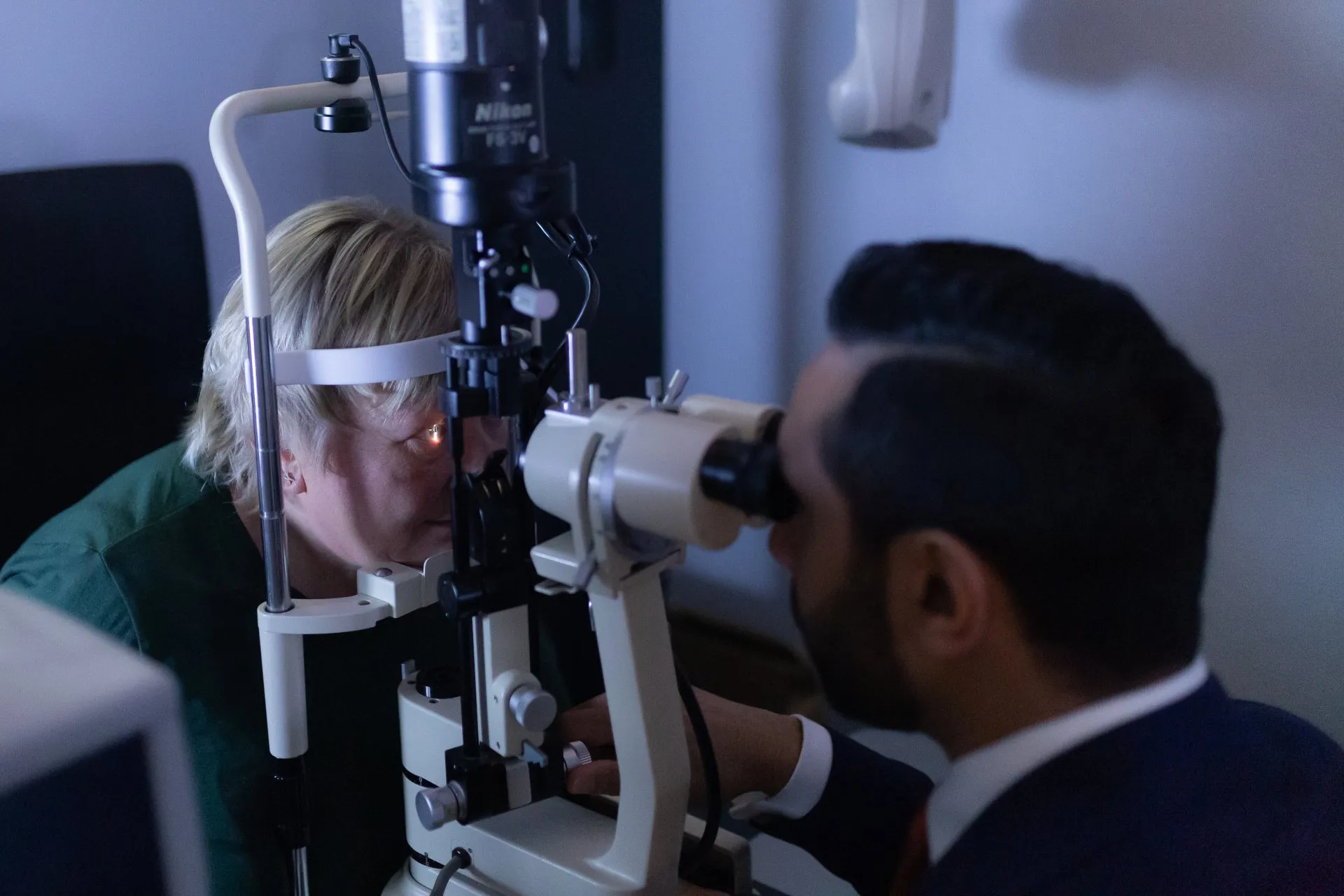 Optometrist conducting eye examination using slit lamp with patient in green shirt