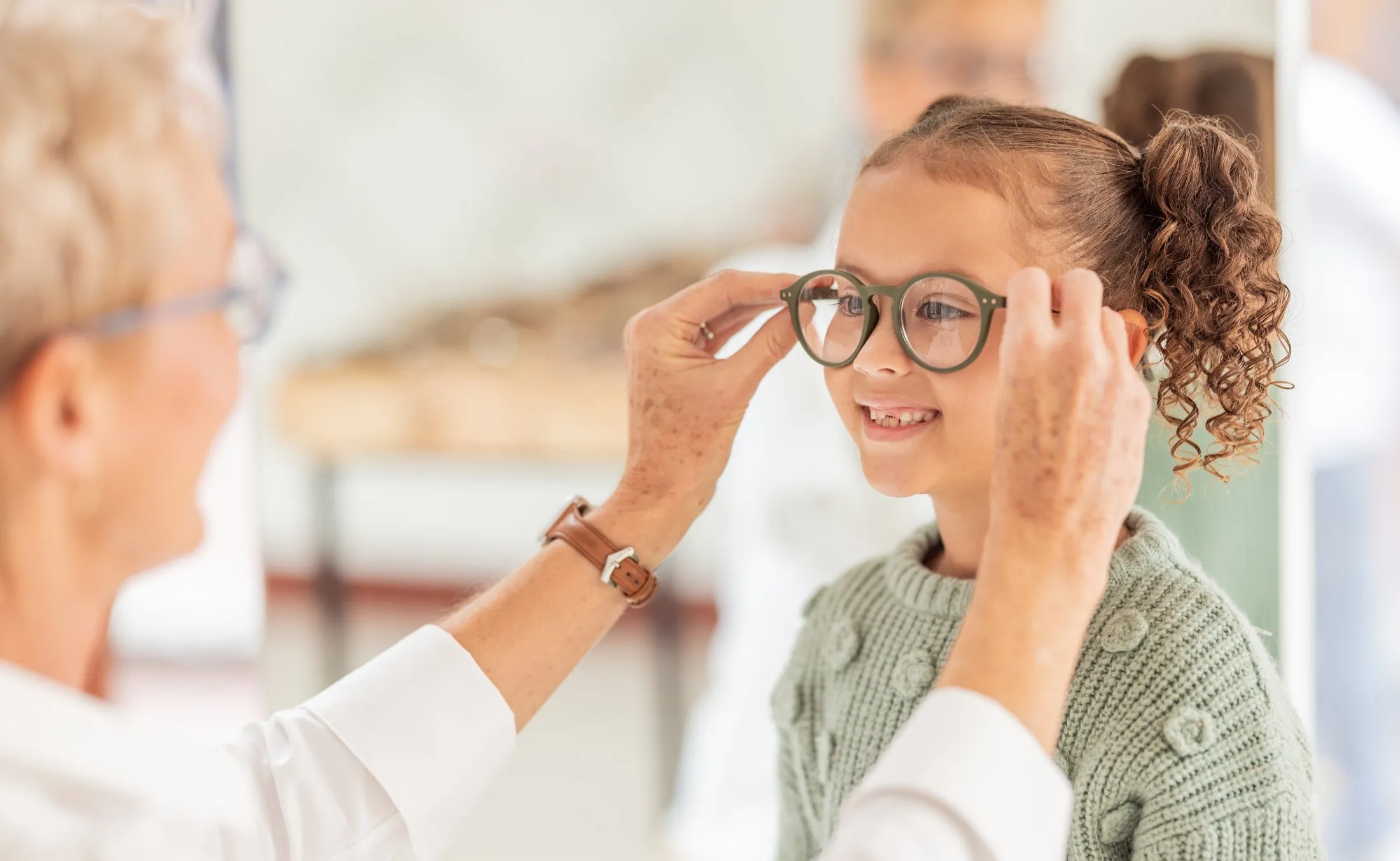 Optometrist fitting eyeglasses on smiling young girl at eye exam