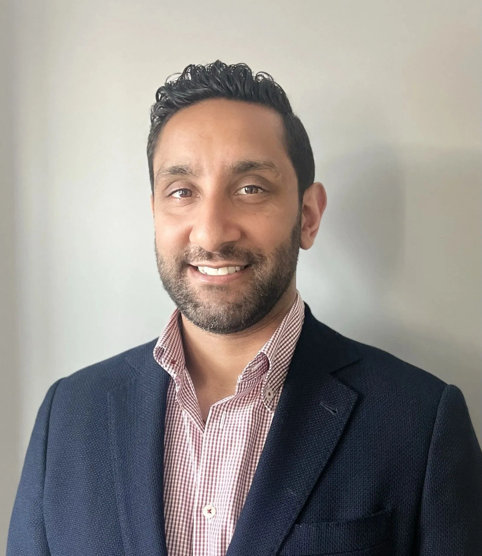 Professional portrait of a smiling man with dark hair, beard, wearing navy blazer and checkered shirt.