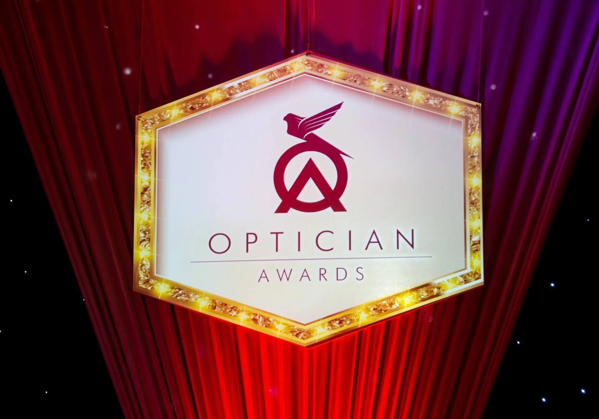 Optician Awards logo on gold-framed hexagon sign against red and purple curtains