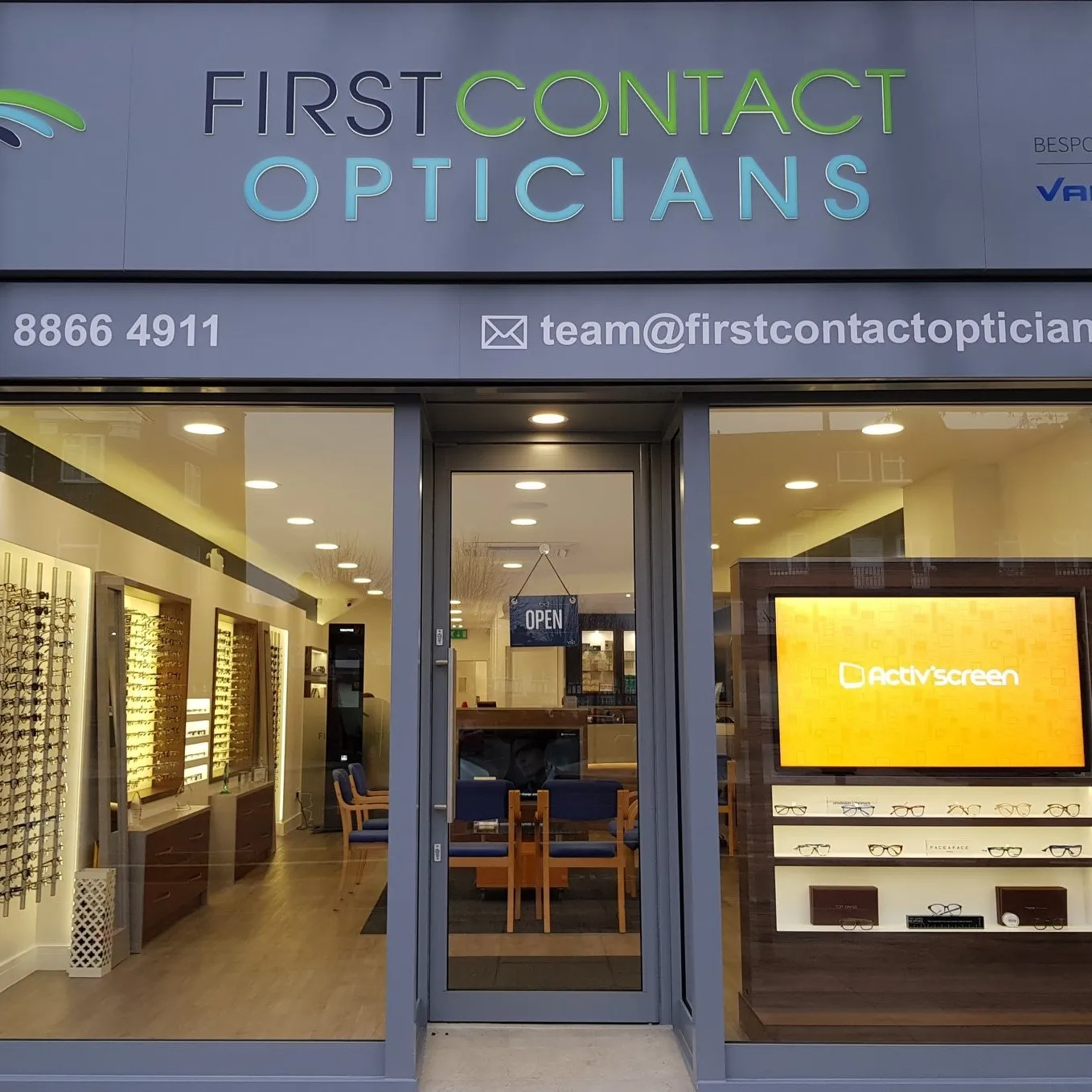 First Contact Opticians storefront with illuminated sign, open door, and eyewear displays inside