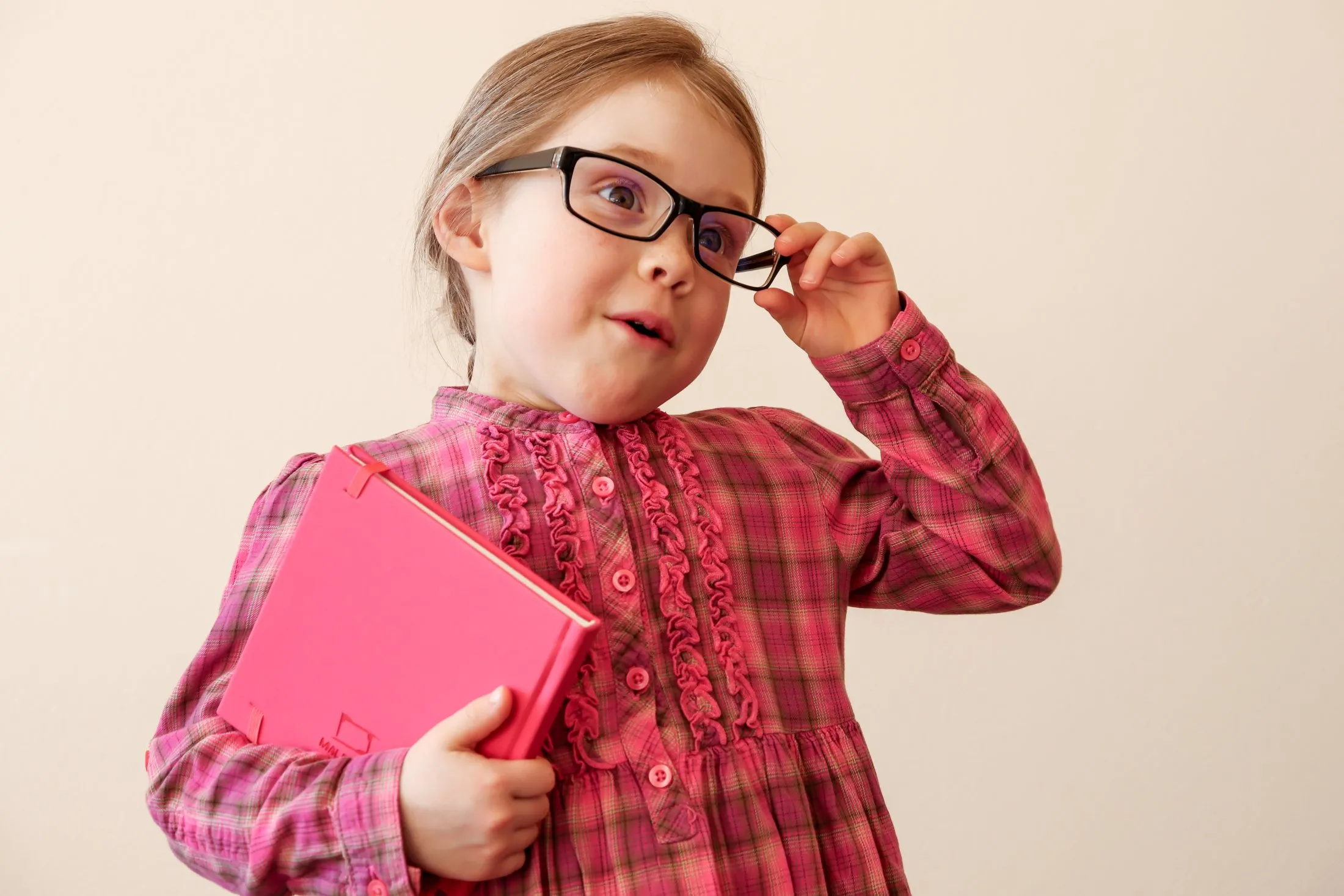 Young girl wearing glasses holding pink book looking upward thoughtfully